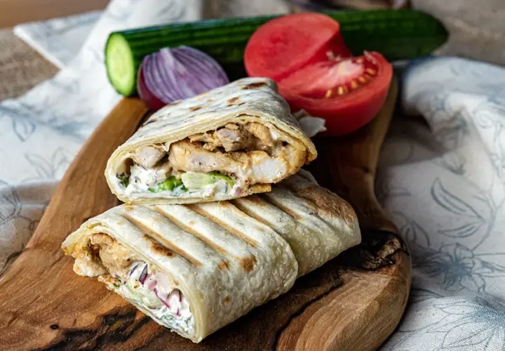 Pyszny kebab w tortilli z kurczakiem, warzywami i sosem, podany na desce z pomidorem i ogórkiem.
