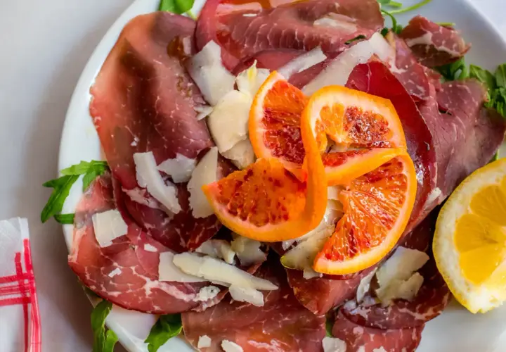 Wykwintna szynka bresaola z płatkami parmezanu, krwistymi pomarańczami i cytryną na talerzu.