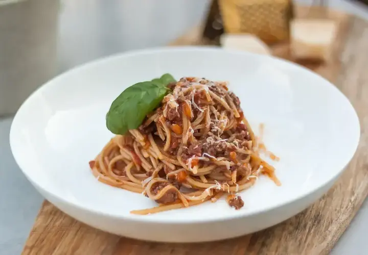 Porcja spaghetti bolognese z parmezanem i listkiem bazylii na białym talerzu.