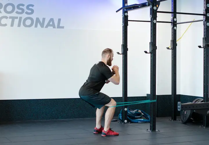 Mężczyzna wykonuje ćwiczenie spanish squat z gumą oporową wokół ud, w siłowni.