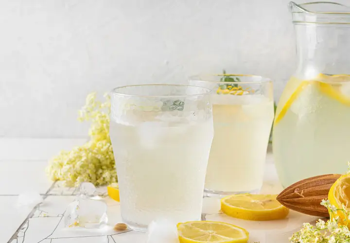 Deux verres de citronnade maison rafraîchissante avec des glaçons, des rondelles de citron, des fleurs de sureau et une carafe remplie.