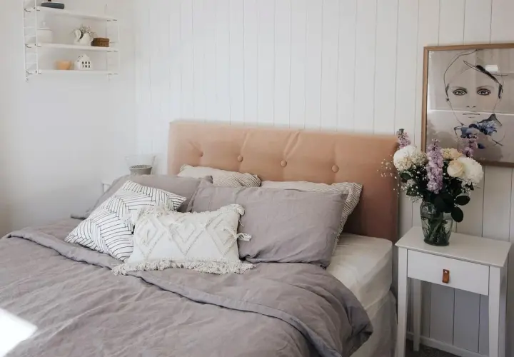 A cozy bedroom featuring a DIY upholstered headboard in a soft peach velvet, adorned with tufted buttons. The bed is dressed in linen bedding and an assortment of decorative pillows.