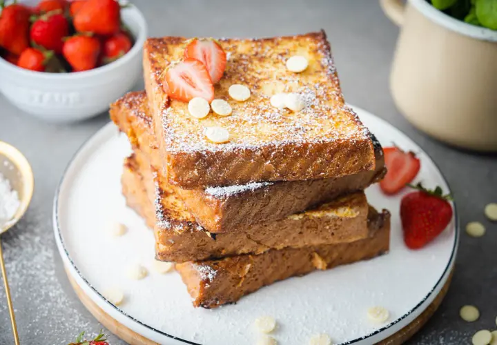 Gestapelte French Toast Scheiben, bestäubt mit Puderzucker und garniert mit Erdbeeren und weißen Schokoladenchips.