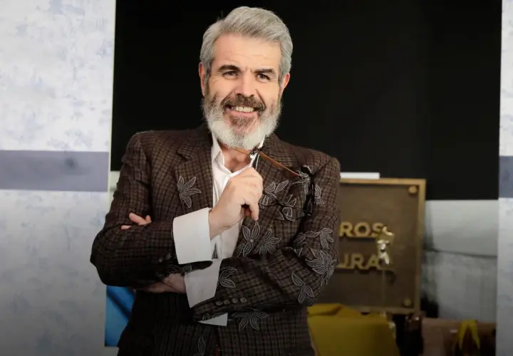 Lorenzo Caprile, con su característica barba canosa, sonríe mientras sostiene unas gafas. Viste una chaqueta de tweed con bordados florales.