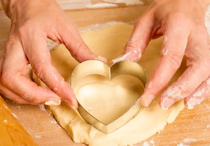 Manos cortando masa con un cortador en forma de corazón, parte del proceso de como hacer galletas caseras.