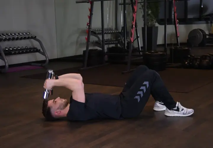 Mężczyzna wykonuje francuskie wyciskanie hantli leżąc na podłodze. Ćwiczenie na triceps.