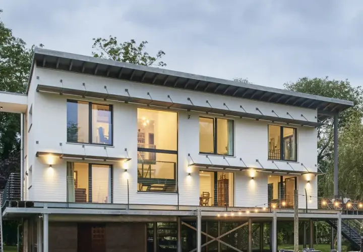 Modernes Haus auf Stelzen mit großen Fenstern und Metalltreppen, umgeben von Bäumen. Lichterketten beleuchten die Terrasse.