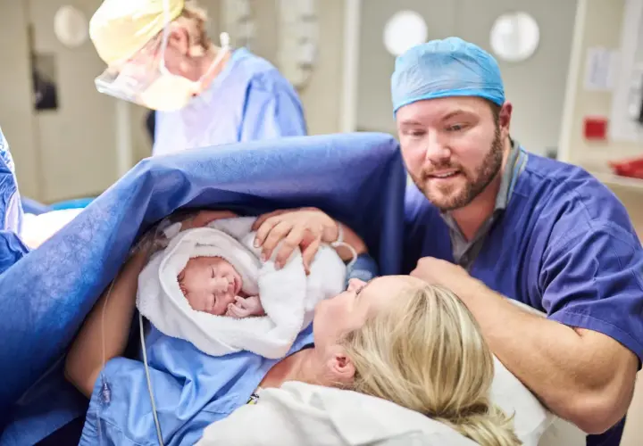 Un padre guarda con amore il suo neonato, avvolto in un asciugamano bianco, dopo un cesareo dolce. La madre sorride, circondata da personale medico.