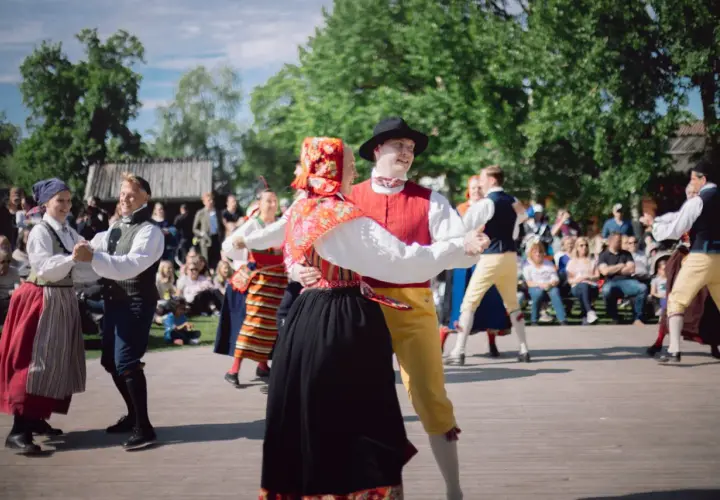 Dansande par i folkdräkter på en utescen, publiken tittar på. En härlig konsert på Skansen.