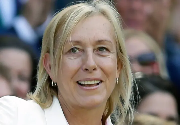 Uśmiechnięta Martina Navratilova w białej marynarce, z blond włosami i kolczykami w kształcie kółek, obserwuje wydarzenie.