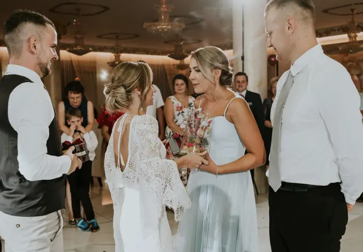 Nietypowe podziękowania dla rodziców na weselu: panna młoda w koronkowej sukni wręcza bukiet w szkle, a pan młody prezent z wstążką.