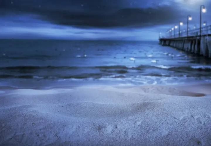 Nocna, świecąca plaża z molo i latarniami w oddali. Piasek lśni w księżycowym blasku.