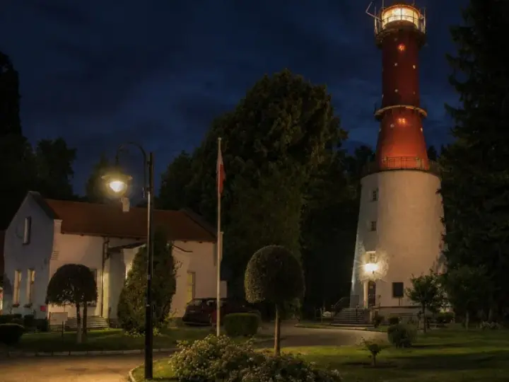 Nocą rozświetlona latarnia morska Rozewie strzeże wybrzeża. Obok niej stoją budynki i ceglany komin.