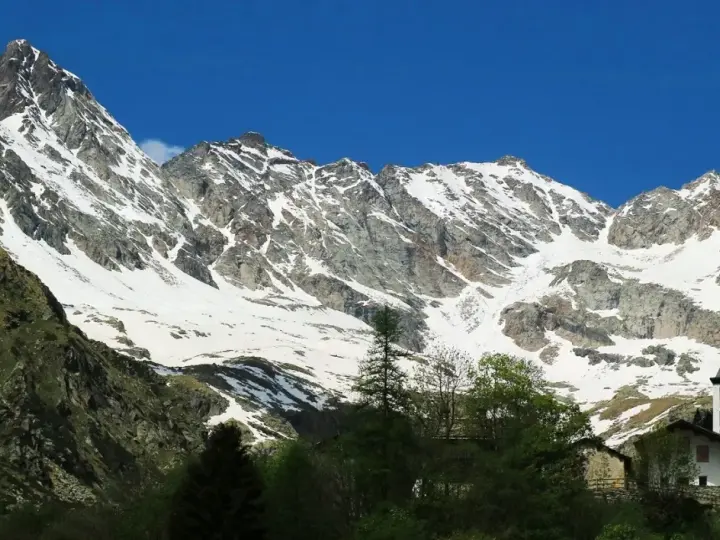 Imponente montagna sacra innevata, con un piccolo campanile bianco che emerge tra gli alberi verdi in primo piano.