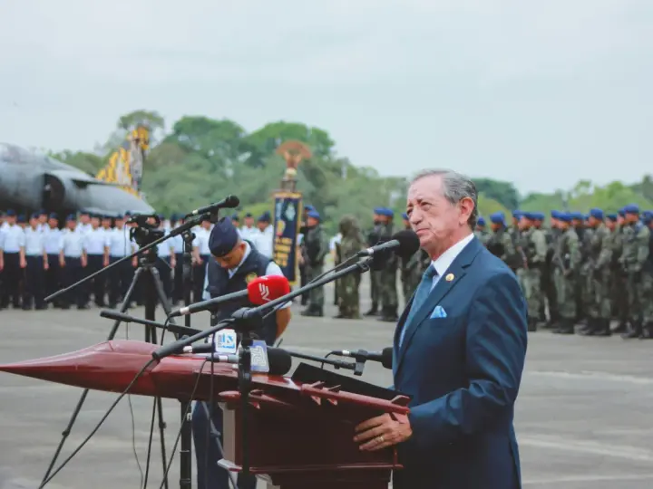 Elegancki garnitur to dobry wybór, gdy zastanawiasz się, jak się ubrać na przysięgę wojskową. Mężczyzna w granatowym garniturze przemawia na tle żołnierzy i samolotu.