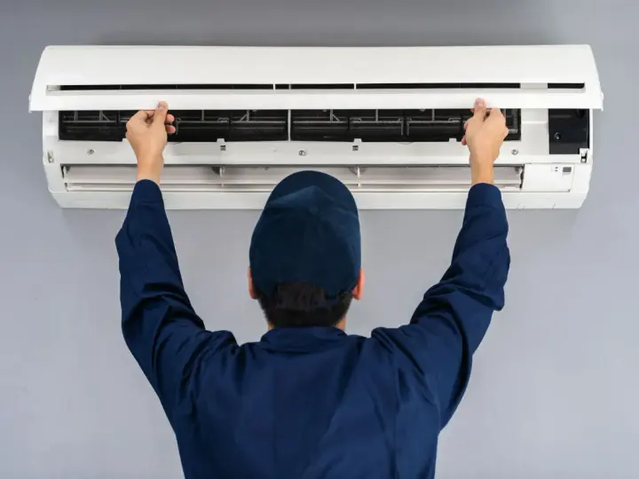 Tecnico con cappellino blu apre il pannello di un climatizzatore per impostare aria fredda.