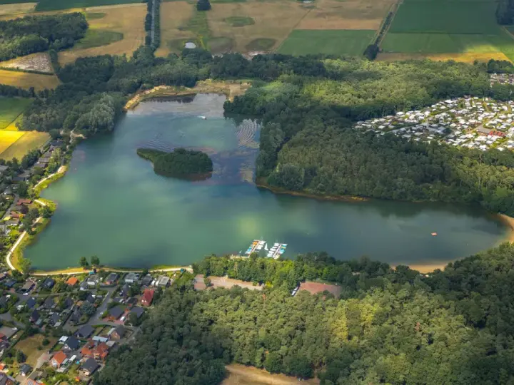 Luftaufnahme des Feldmarksees Sassenberg mit einer kleinen Insel, Booten am Steg, einem Wohngebiet und Feldern.