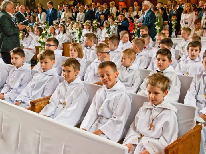 Dzieci w bieli, gotowe na rocznicę komunii świętej, siedzą w kościele. W tle widać zgromadzonych gości i fotografa.