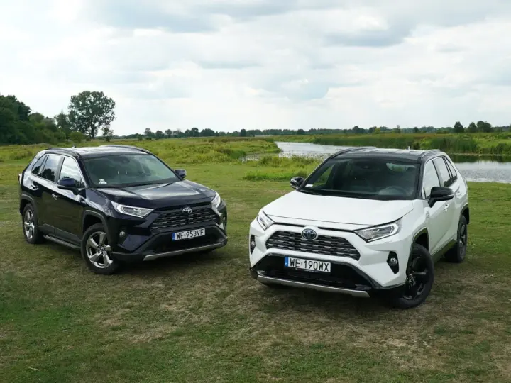 Dwa SUV-y: ciemny i biały Toyota RAV4 hybryda, stoją na łące nad jeziorem.