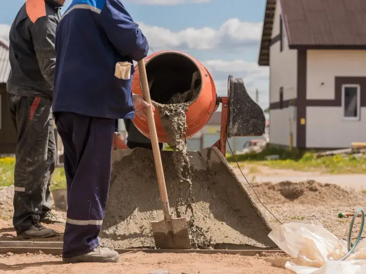 Dwóch budowlańców miesza beton w betoniarce. Widać, ile żwiru na worek cementu potrzeba do uzyskania odpowiedniej konsystencji.