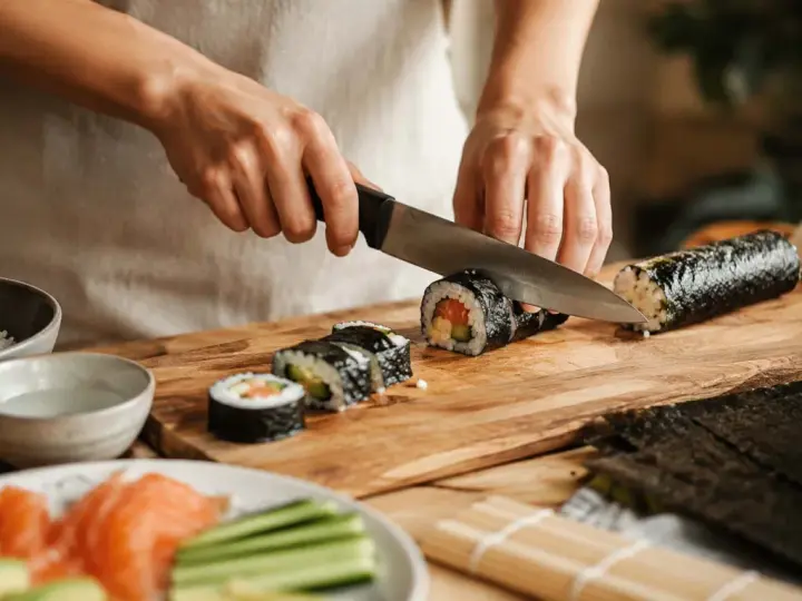 Oto jak się robi sushi: ręce kroją rolkę sushi na desce. Obok ryż, łosoś, awokado i ogórek.
