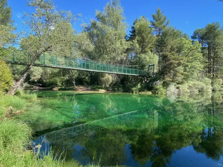 Puente verde cruza un río de aguas cristalinas y verdes, reflejando el bosque exuberante. Un lugar idílico para explorar el **alto tajo**.