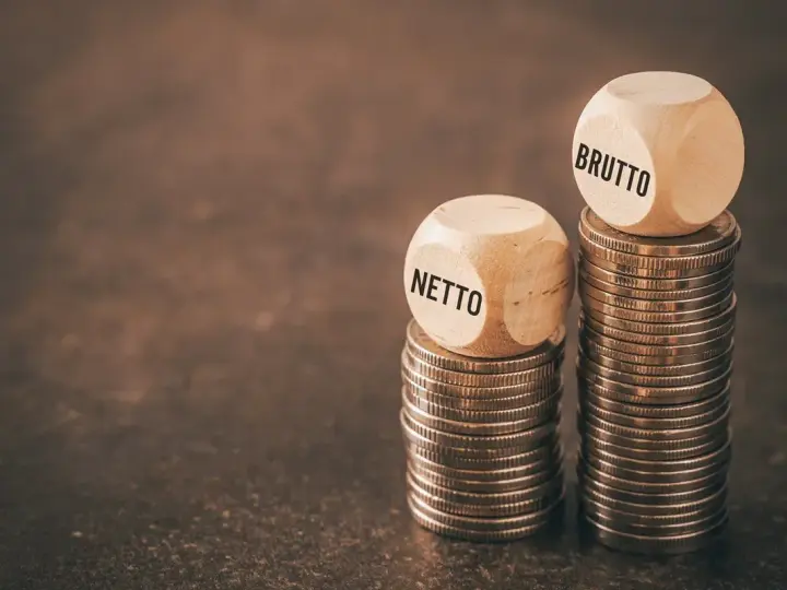 Dwie wieże monet, jedna z kostką "NETTO", druga z "BRUTTO". Symbolizuje to rozważania nad umową zlecenie i stawką godzinową.