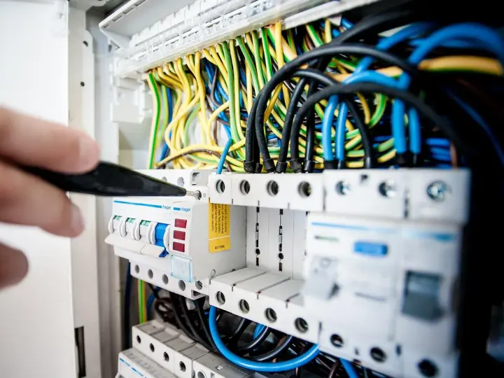 Ręka majstruje przy bezpiecznikach. Kolorowe przewody tworzą skomplikowaną instalację elektryczną.