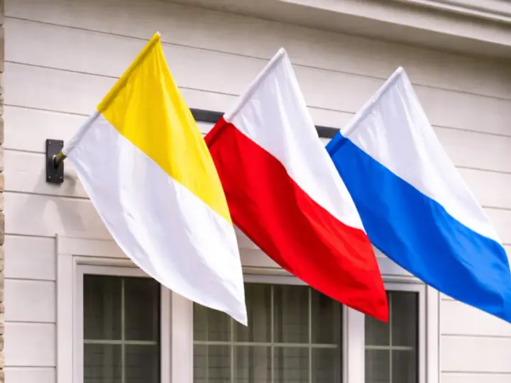Trzy flagi: żółto-biała, czerwono-biała i niebiesko-biała, powiewają na wietrze. To może być flaga kościelna lub symbol parafii.