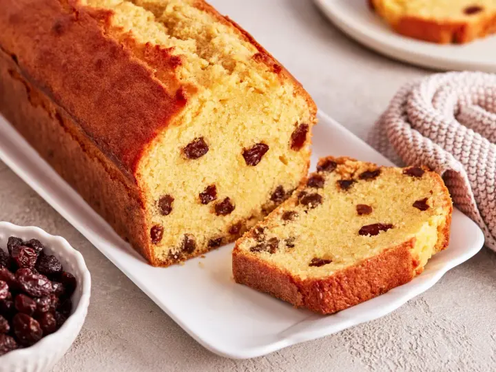 Ein saftiger Rosinenkuchen mit goldbrauner Kruste, auf einem weißen Teller angerichtet. Rosinen sind gut sichtbar im Teig verteilt.