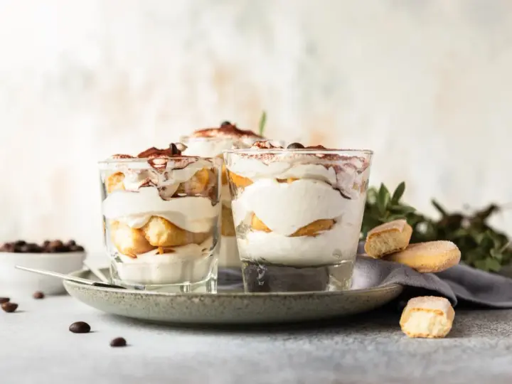 Pyszne desery włoskie: tiramisu w szklankach, udekorowane kakao i kawą. Obok kawałki biszkoptów i ziarna kawy.