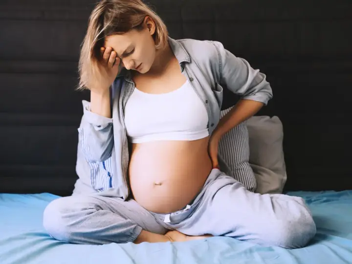 Ciężarna kobieta siedzi na łóżku, trzymając się za głowę i plecy. Odczuwa ból krocza w ciąży, co widać po jej wyrazie twarzy.