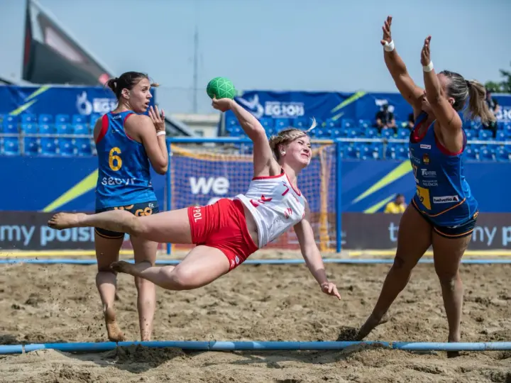 Piłkarka w locie rzuca zieloną piłką podczas meczu beach handball. Rywalki próbują ją zatrzymać.