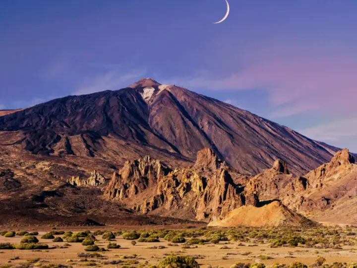 Krajobraz w parku narodowym Teide z księżycem w kształcie sierpa na fioletowym niebie. Wulkan góruje nad skalistym terenem.