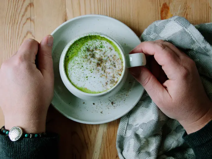 Händerna håller en kopp med en krämig matcha latte, toppad med skum och lite kryddor. En mysig stund med en grön dryck.