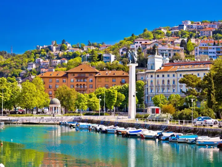 Malowniczy port w Chorwacji z łodziami przycumowanymi do nabrzeża. W tle widać wzgórze z zabudowaniami i zamek.