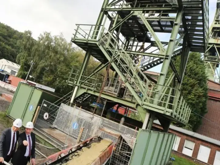 Drei Männer in Anzügen und Schutzhelmen inspizieren das Gelände der Zeche Bottrop. Ein Förderturm ragt in den Himmel.