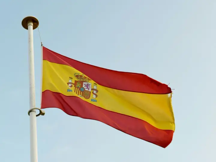Spanska flaggan vajar i vinden. Den har tre horisontella band: rött, gult och rött. I det gula bandet finns Spaniens riksvapen. Orden "spanska ord" kan associeras med denna flagga.