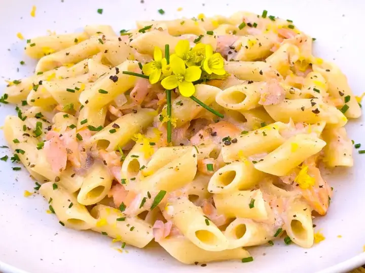 Pasta penne cremosa con trozos de salmón rosado, ralladura de limón y flores amarillas.