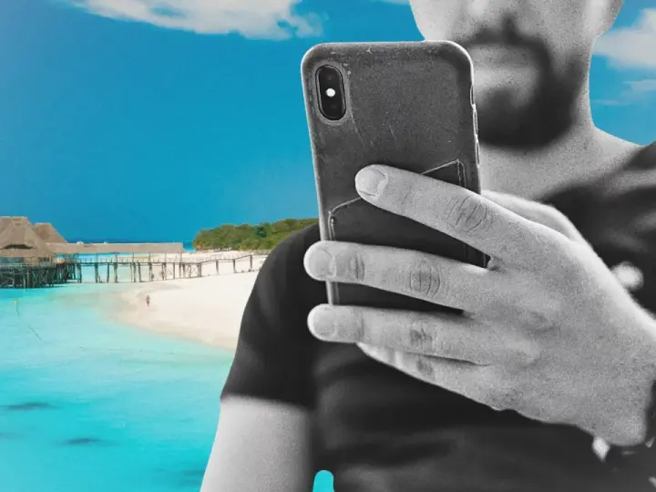 Mężczyzna z telefonem na plaży na Zanzibarze, sprawdzający zanzibar internet. W tle turkusowa woda i drewniany pomost.