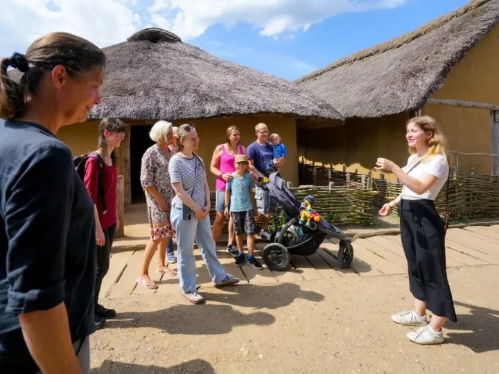 Familienurlaub in Haithabu: Eine Gruppe erkundet historische Häuser mit Strohdächern, während eine junge Frau eine Führung gibt.