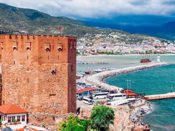 Alanya zwiedzanie na własną rękę: potężna czerwona wieża zamku, port z łodziami, latarnia morska i miasto w tle.