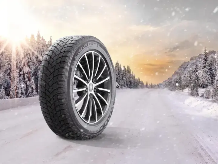 Michelin X-Ice Snow-däck på en snötäckt väg i en vinterskog. Solen bryter igenom molnen.