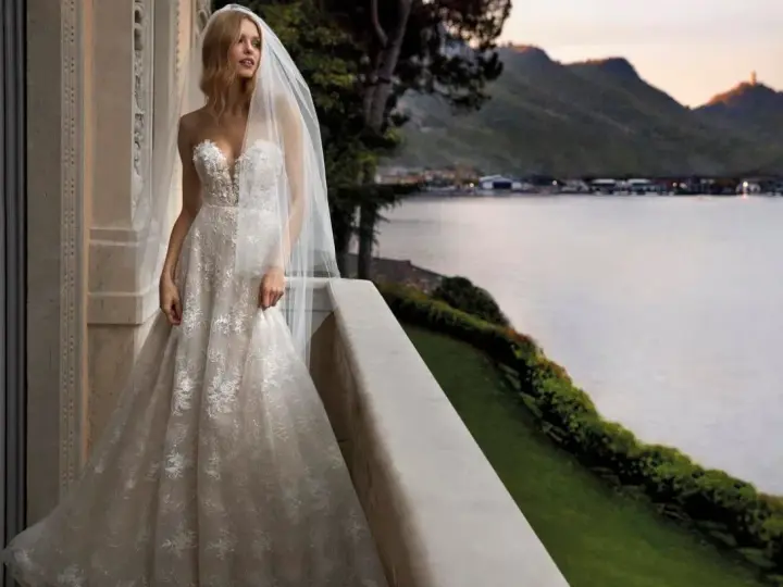 Sposa con abito da sogno, con vari tipi di pizzo che scendono a cascata, su un balcone con vista lago al tramonto.