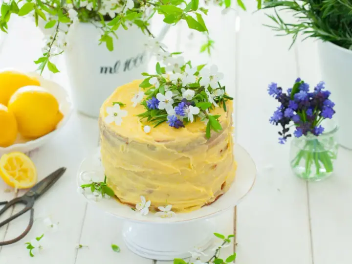 Słoneczny tort cytrynowy z kremem lemon curd, ozdobiony świeżymi kwiatami i ziołami. Obok leżą cytryny i stare nożyczki.