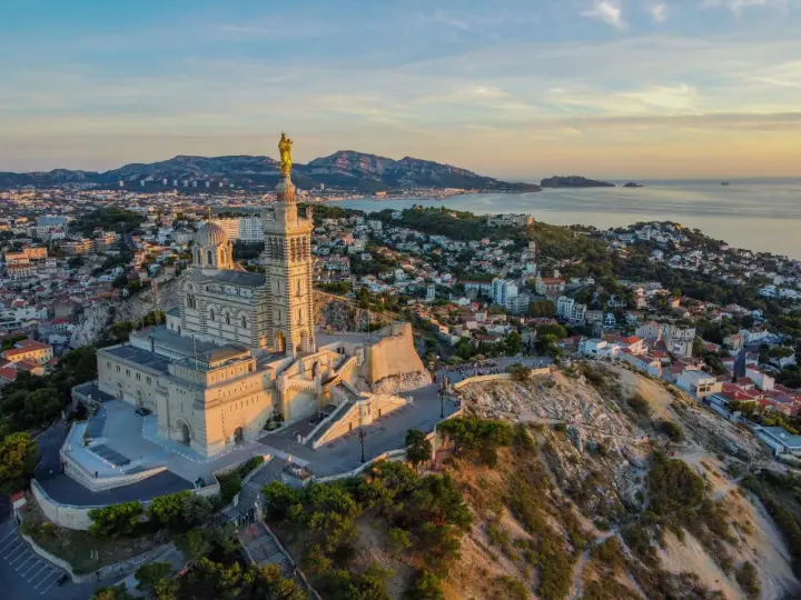 La basilique Notre-Dame de la Garde veille sur Marseille, baignée par la lumière dorée du soleil couchant. La mer et les collines environnantes s'étendent à perte de vue.