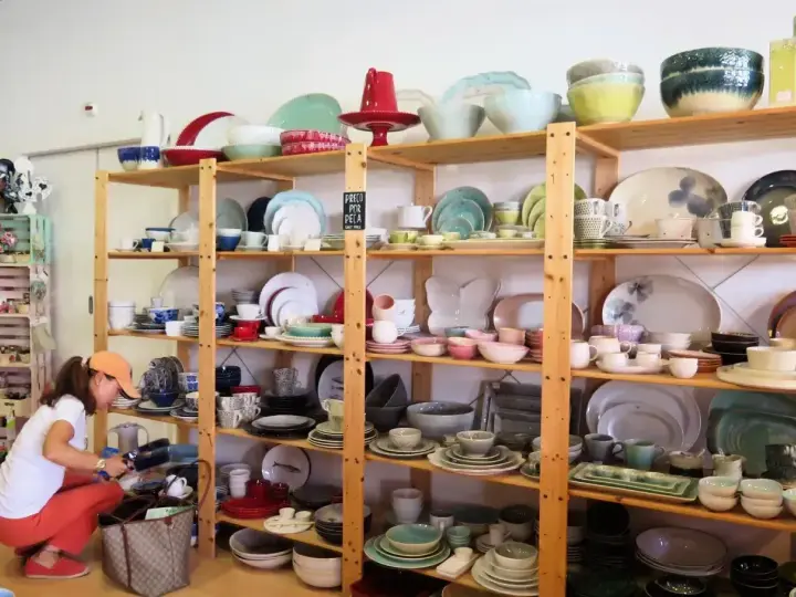 Kobieta wybiera ceramiczne naczynia, idealne pamiątki, które warto co przywieźć z Portugalii. Kolorowe talerze, miski i filiżanki na drewnianych półkach.