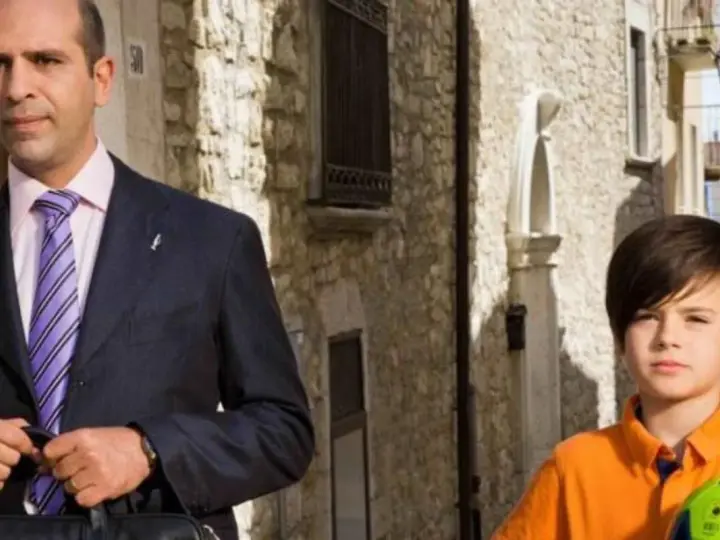 Checco Zalone con uno dei suoi figli, un bambino con un pallone da calcio, passeggiano in un vicolo.