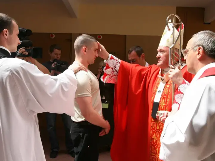 Biskup udziela sakramentu bierzmowania młodemu mężczyźnie. W tle widać osoby z kamerami.