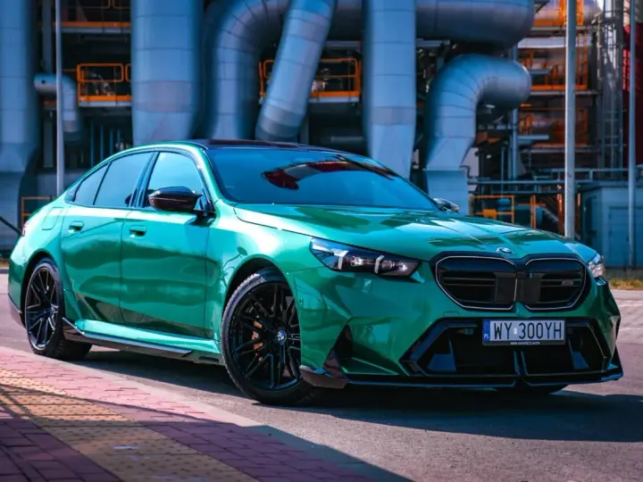 Błyszczące, zielone BMW M5 G90 z czarnymi felgami stoi na tle przemysłowych rur.